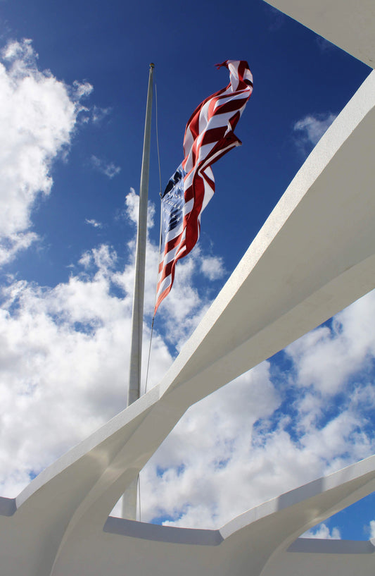 An image of the American flag waving in the wind at Pearl Harbor, with the flag prominently displayed against a blue sky with clouds, framed by the Memorial's beams.