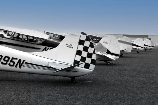 A line of five Cessna plane tails in black and white, with a checkered pattern on the vertical stabilizers, parked on grass under a blue sky. Black and White Cessnas Under Blue Sky, Original Photograph by Kim A. Bailey