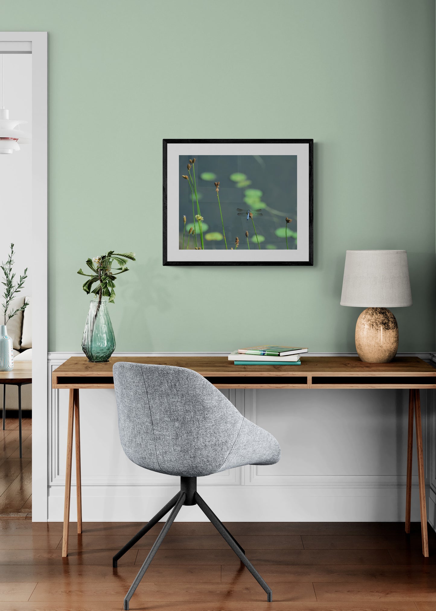 Modern home office with a desk, chair, lamp, and framed artwork on a green wall.