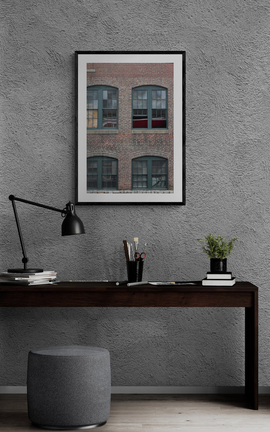 Framed photograph of a brick building above a desk with a lamp and plant.