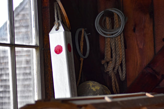 A photograph of a wooden buoy with red dots, hanging on a wall alongside rope and other nautical items, with a window in the background.