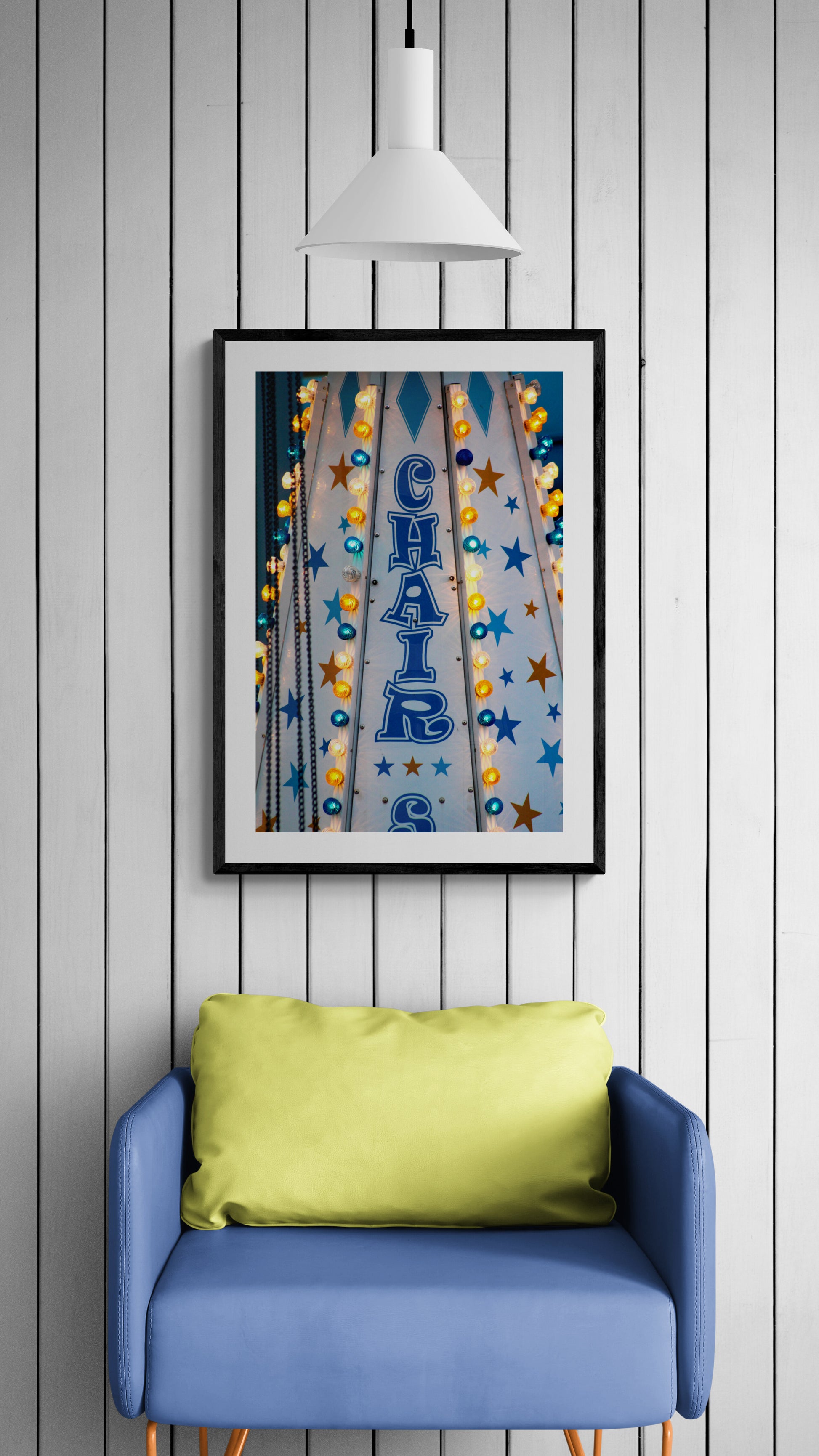 Framed artwork with colorful design above a blue chair with a green pillow against a gray wooden wall.