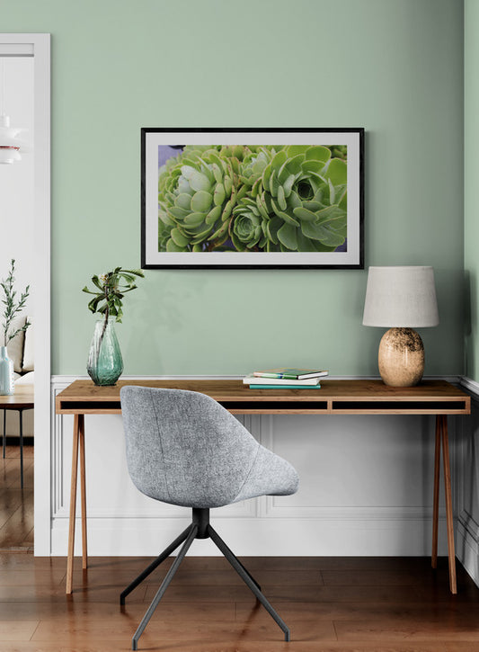 Home office with a desk, chair, lamp, and framed picture of green succulents on a light green wall.