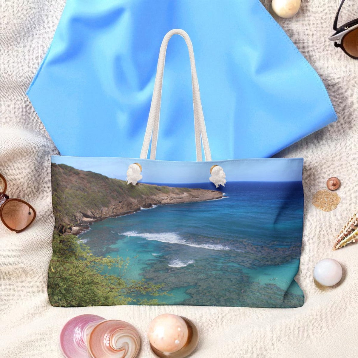 Blue tote bag with a scenic print of a coastal view, surrounded by seashells and sunglasses on a sandy background.