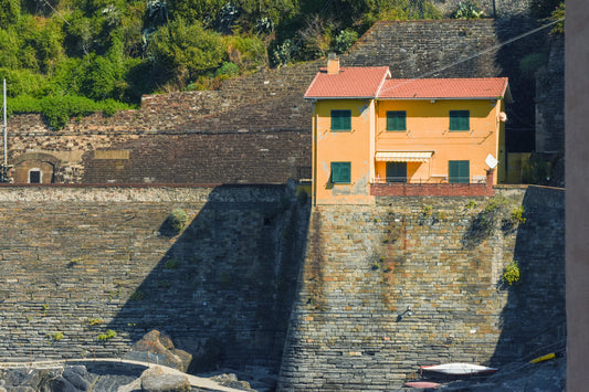 A photograph titled 'House Above the Bay' showing an orange house perched on a rock wall with a beautiful bay and mountains in the background. House Above the Bay, Original Photograph by Kim A. Bailey