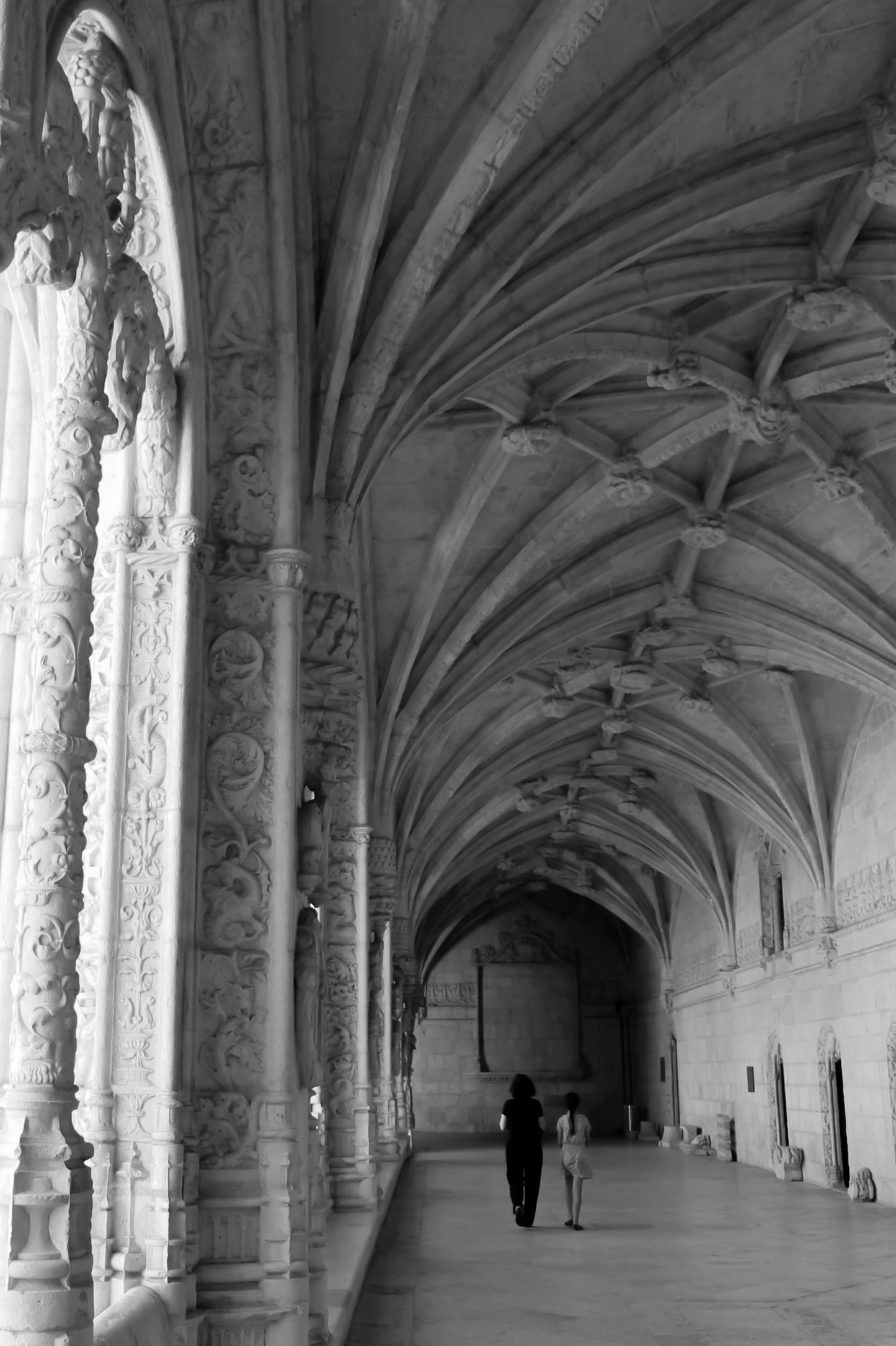 Black and white photograph of the interior of Jeronimos Monastery, Lisbon, Portugal, featuring intricate architectural details and a pair of visitors. Jeronimos Monastery Lisbon, Portugal (B/W) Original Photograph by Kim A. Bailey