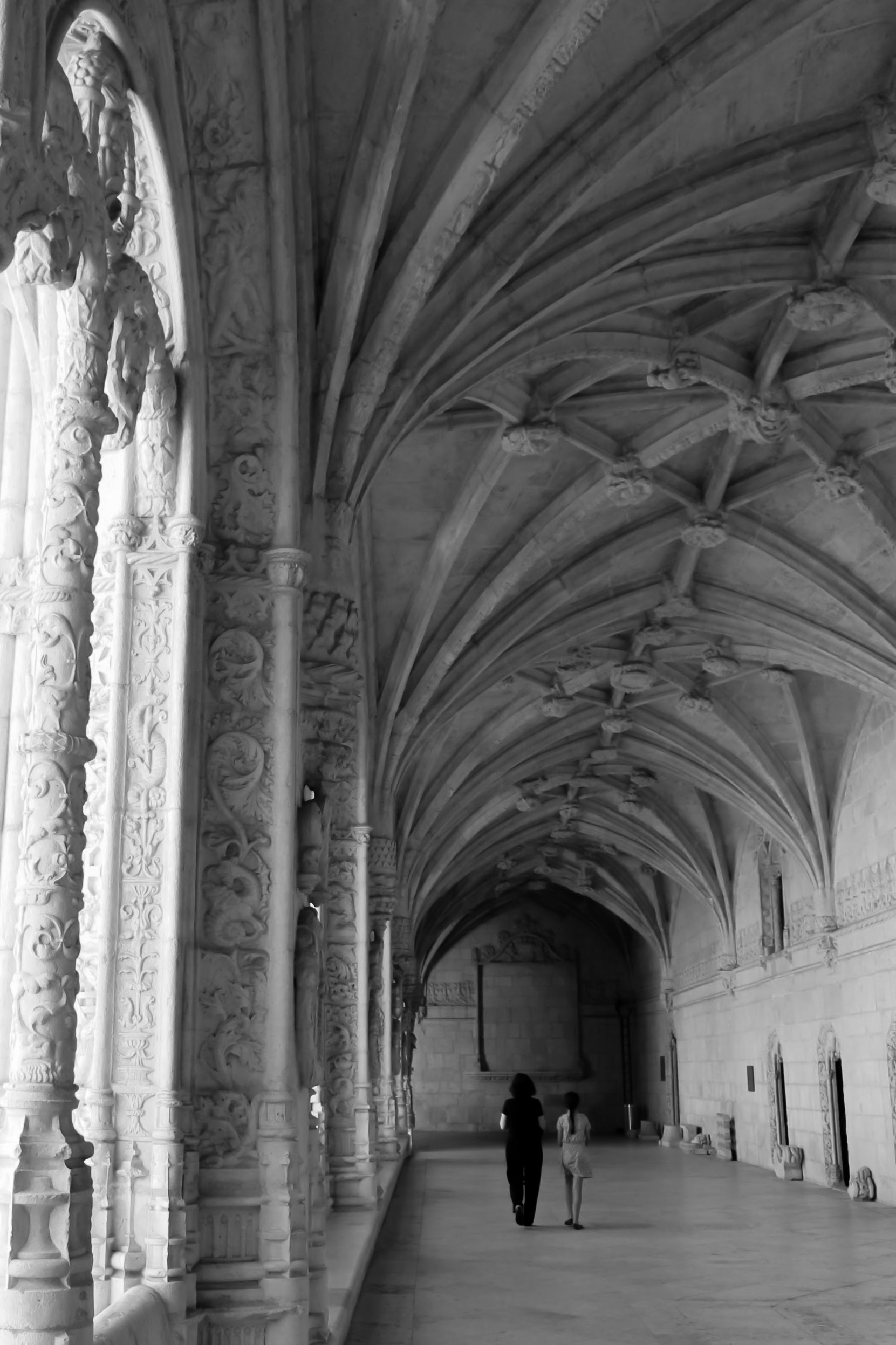 Black and white photograph of the interior of Jeronimos Monastery, Lisbon, Portugal, featuring intricate architectural details and a pair of visitors. Jeronimos Monastery Lisbon, Portugal (B/W) Original Photograph by Kim A. Bailey
