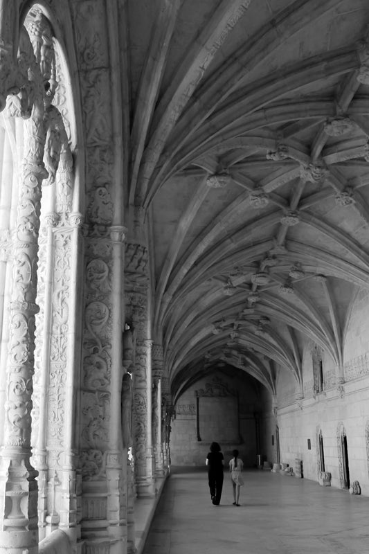 Black and white photograph of the interior of Jeronimos Monastery, Lisbon, Portugal, featuring intricate architectural details and a pair of visitors. Jeronimos Monastery Lisbon, Portugal (B/W) Original Photograph by Kim A. Bailey