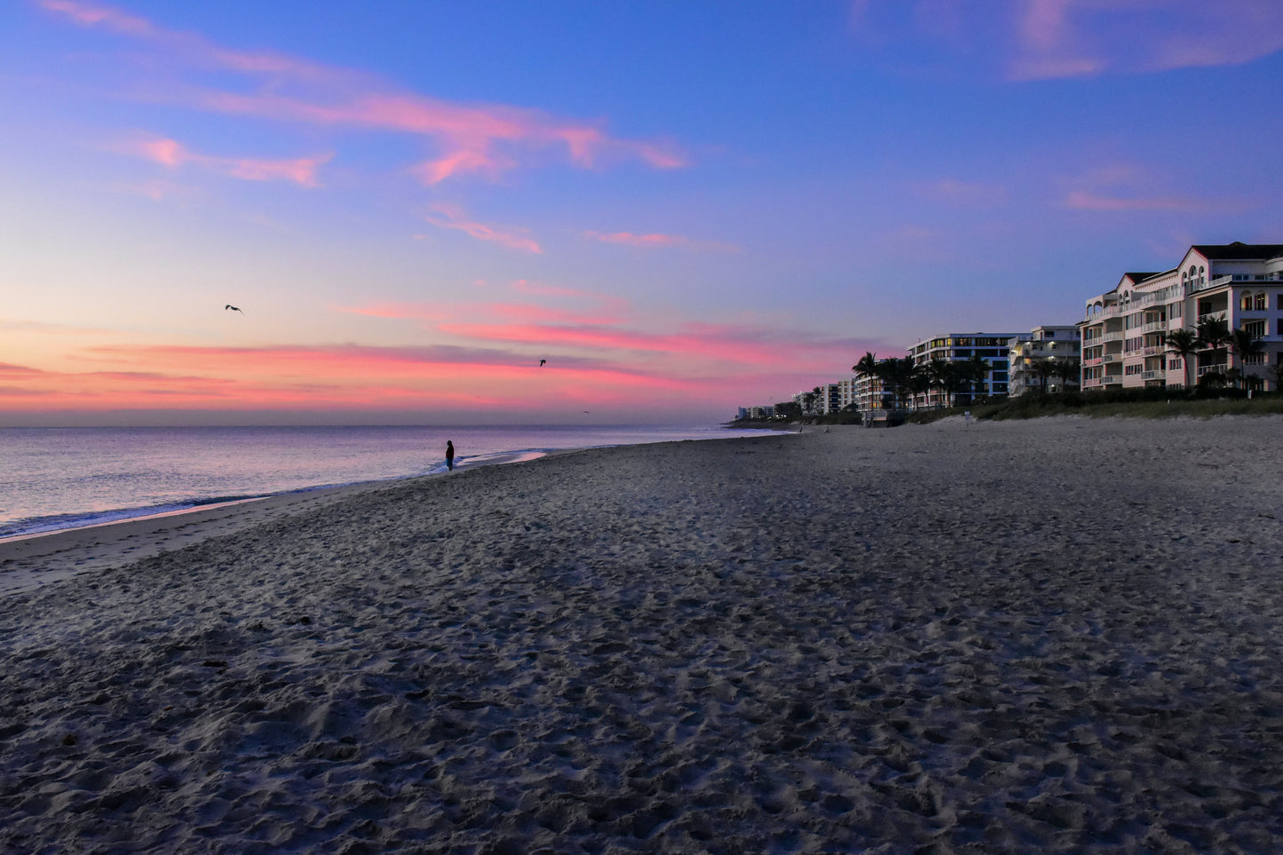 A vibrant image of a sunrise over Lake Worth Beach, Florida, showcasing the colors pink and orange in the sky, reflecting on the water, with silhouettes of people and a beach house in the distance.