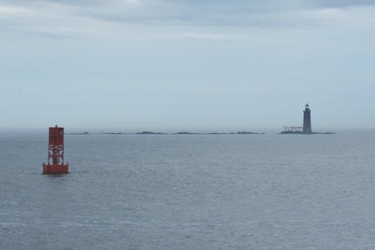 A color photograph featuring a lighthouse and a buoy in the ocean, with a body of water and a cloudy sky in the background Lighthouse and Buoy. Original Photograph by Kim A. Bailey
