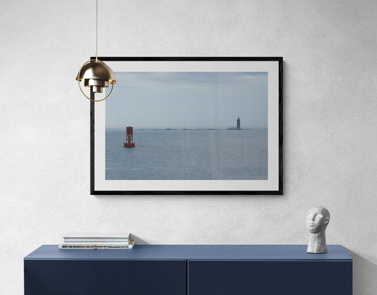 Framed photograph of a lighthouse on the water, above a blue cabinet with a lamp and sculpture. Lighthouse and Buoy Original Photograph by Kim A. Bailey