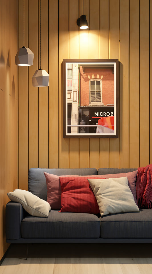 A framed art print depicting a brick building with a window and the text 'MICROB' is displayed on a warm wood-paneled wall above a blue-gray sofa with red and beige pillows.