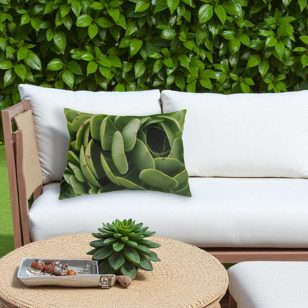 Succulent-patterned pillow on a white outdoor sofa with a green plant and small tray on a table.