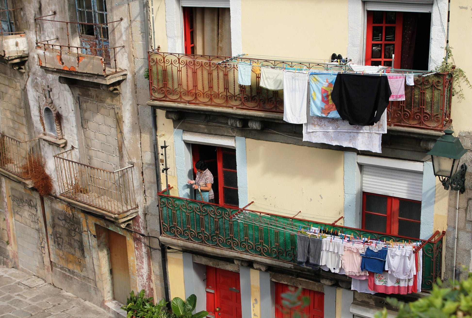 A color photograph depicting a woman hanging laundry on a clothesline in front of a building with balconies and colorful doors in Portugal. Portuguese Woman Hanging Laundry, Original Photograph by Kim A. Bailey