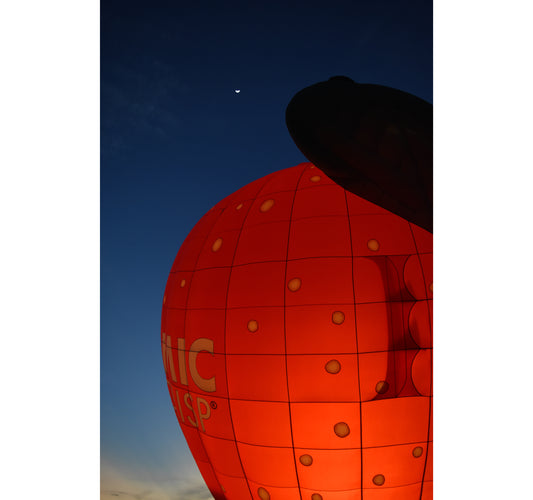 A photograph of a red hot air balloon in the foreground with a clear blue night sky and a crescent moon in the background, taken during the 2022 Lakeland, Florida Introducing Balloon Fest. Red Balloon Under the Moon, Original Photograph by Kim A. Bailey