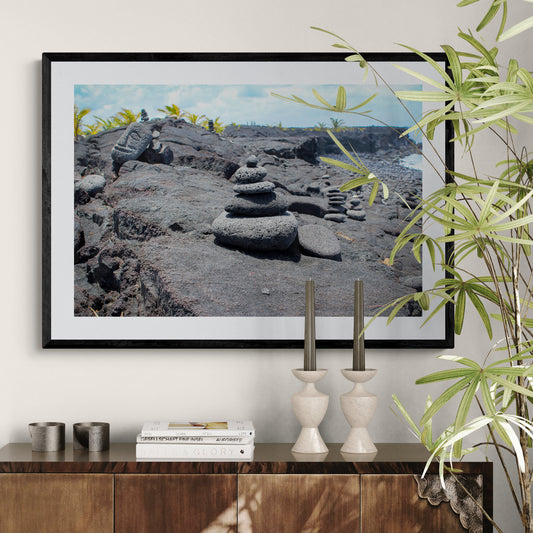 Framed picture of stone pile on rocky shore with plants and candles on table.