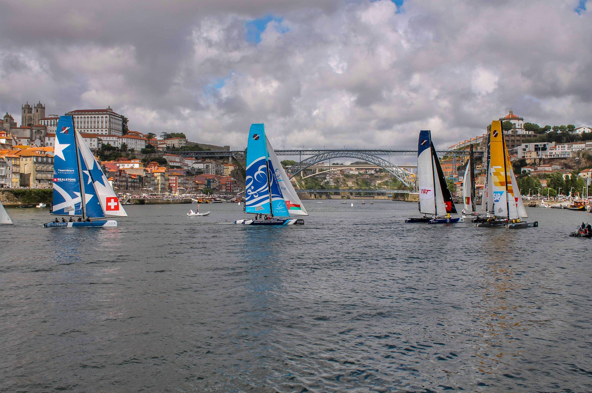 A color photograph depicting a sailing regatta with boats navigating through a narrow channel, accompanied by helicopters, with the city of Porto in the background.