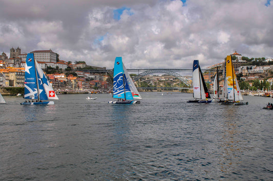A color photograph depicting a sailing regatta with boats navigating through a narrow channel, accompanied by helicopters, with the city of Porto in the background.