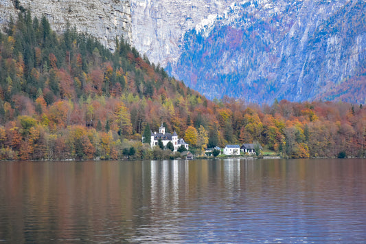 A photograph of Schloss Grub Castle in Austria, surrounded by a lake and a forested landscape in autumn colors, with a dreamlike quality due to the castle's reflection in the still water and a misty haze in the background.