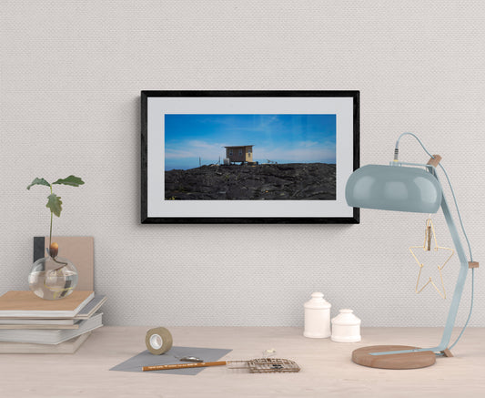 Framed photograph of a small hut on a rocky landscape, displayed on a wall with a lamp and plant.
