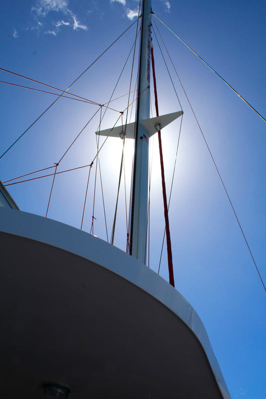 Photograph of a sailing ship's mast silhouetted against a bright sun, with a soft focus on the rigging and a clear blue sky in the background.