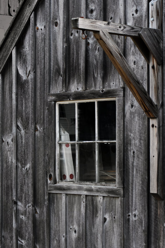 A color photograph featuring a weathered exterior wood shack with a visible window containing a fishing buoy, reflecting a sense of nostalgia and coastal life. Single_Wood_Buoy Original Photograph by Kim A. Bailey