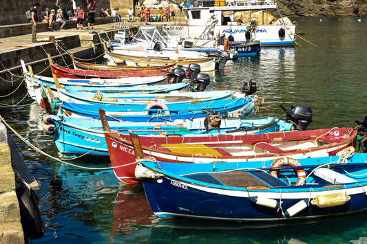A photograph showcasing a group of small, colorful wooden fishing boats gently bobbing in the water, with the Cinque Terre coastline in the background. Small Boats in the Bay, Original Photograph by Kim A. Bailey