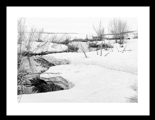 A black and white photograph of a serene winter scene with snow on the ground and a partially frozen creek, with footprints or marks on the snow surface.