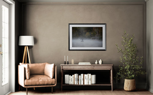 Person on a boat in a foggy lake with a gazebo in the background in a modern living room with a brown armchair, side table, and wall art.