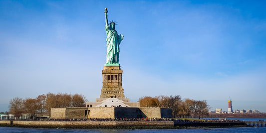 A color photograph of the Statue of Liberty by Kim A. Bailey, showing the statue in clear detail with a blue sky in the background and a crowd at the base.