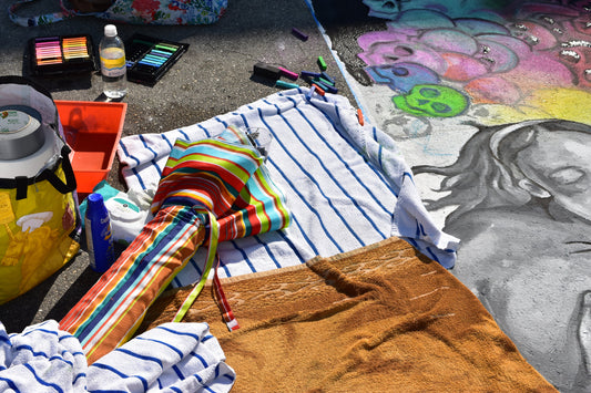 A photograph titled 'Supplies of a Street Painter' showing an array of painting tools and materials including a colorful umbrella, chalks, and canvases laid out on the ground. Supplies of a Street Painter