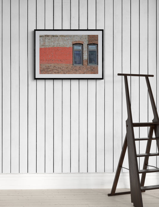 Framed artwork of a brick wall with a window on a white paneled wall.