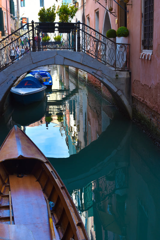 A color photograph of a quiet canal in Venice, Italy, with a boat docked on one side and a bridge overhead, reflecting in the still water.