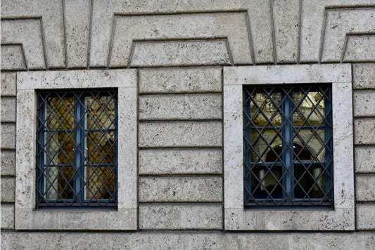 Two windows with geometric concrete framing and decorative grilles, set against a gray stone wall.