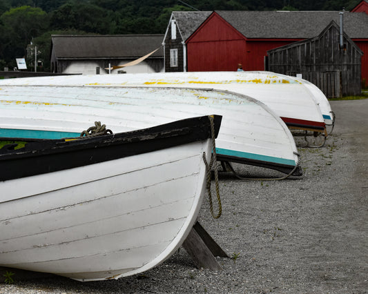 A photograph of classic wooden boats propped on the sand and rock, with quaint red and gray shacks in the background, capturing the essence of Mystic, Connecticut. Wooden Boats Original Photograph by Kim A. Bailey