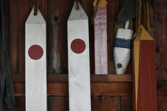 A photograph of wooden fishing buoys with red and blue markings, hanging on a wooden wall, likely in a shack or storage area. Wooden Buoys Original Photograph by Kim A. Bailey