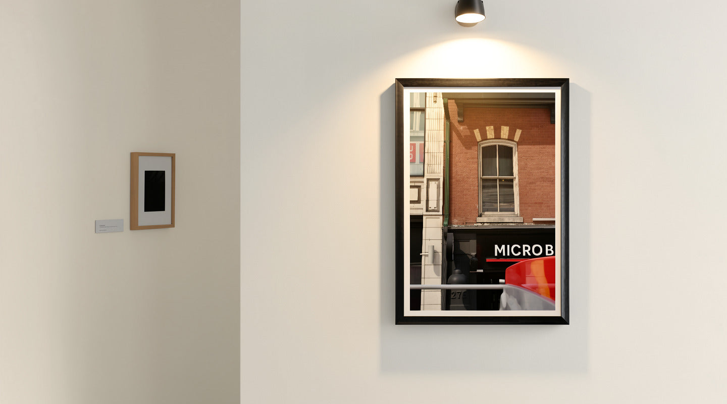 A framed art print featuring a street scene with a brick building and a sign reading 'MICROB' is displayed on a white gallery wall under a spotlight. Another small framed piece hangs to the left.
