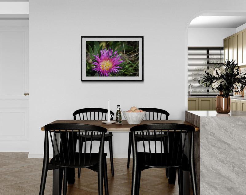 Dining area with a table and chairs, framed floral picture on the wall, and a kitchen in the background.