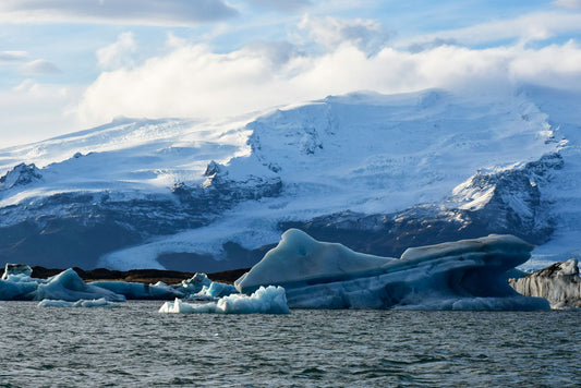 An iceburg floating in a glacial lagoon with a snow-covered mountain in the background, representing the Blue Glacier Ice of Iceland.
