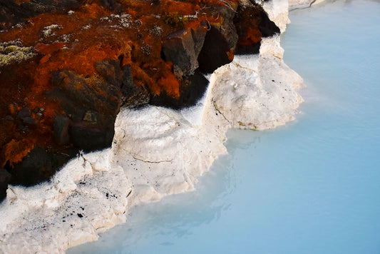 Photograph of the edge of the Blue Lagoon in Iceland, showing the lagoon's blue water next to red lava rock with white mineral deposits.