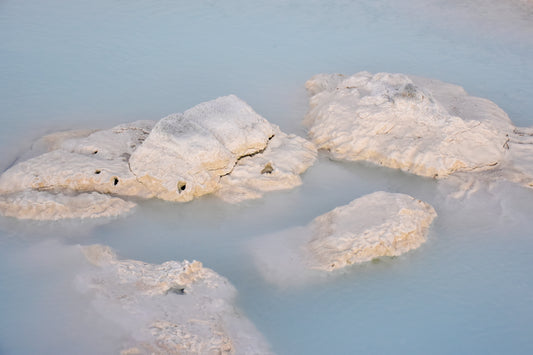 White and light blue rocks partially covered in silicon, situated in a blue body of water, with steam rising from the surface.