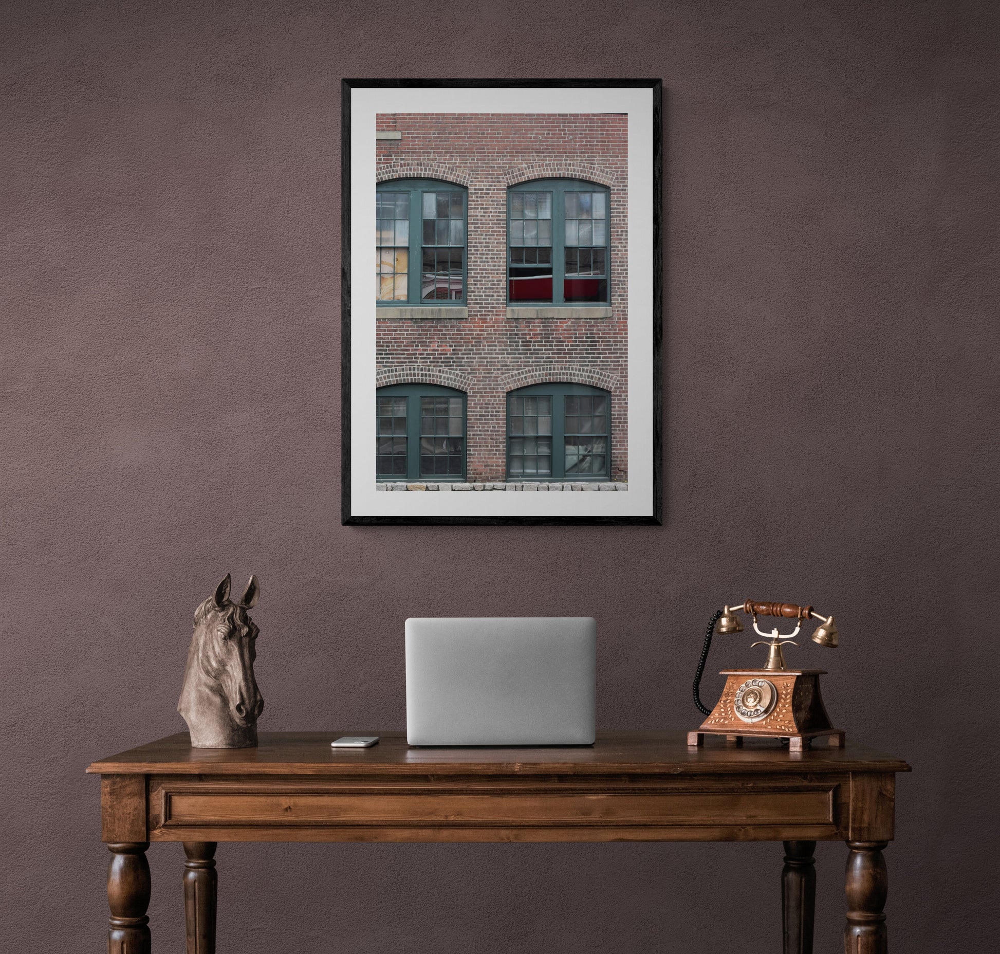 Framed photograph of a brick building above a wooden desk with a laptop and horse statue.