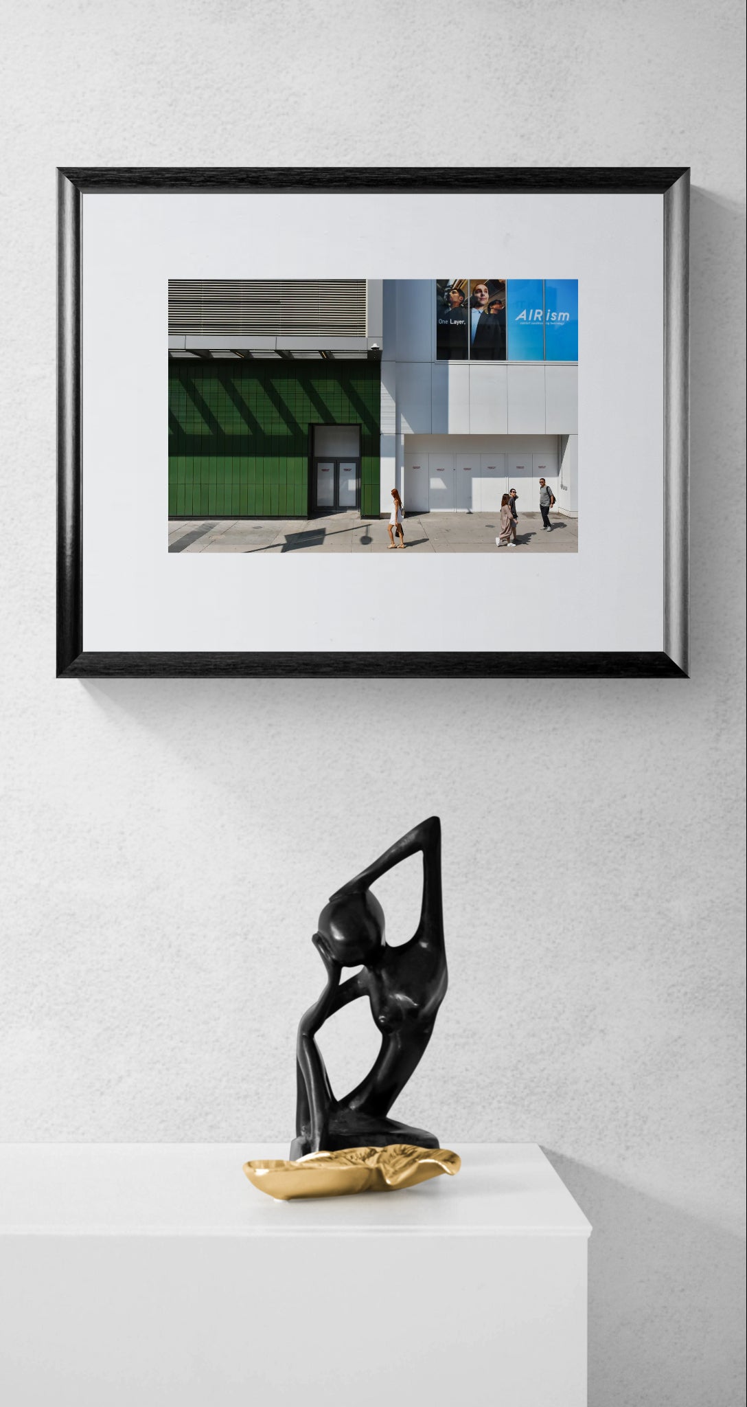 Urban Photograph in big blocks of white, grey, blue, and green colors. People walking through the scene add scale. Framed photograph hanging over a stone sculpture.