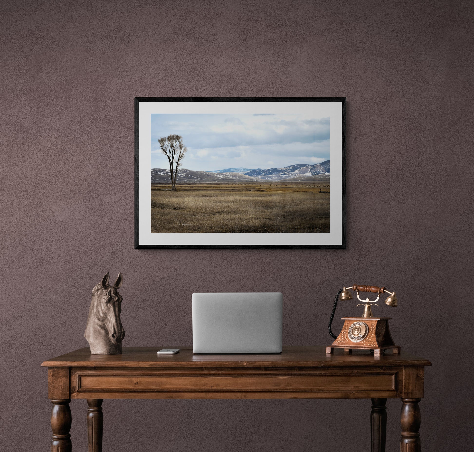 Framed landscape print above a wooden desk with a laptop and decorative items.