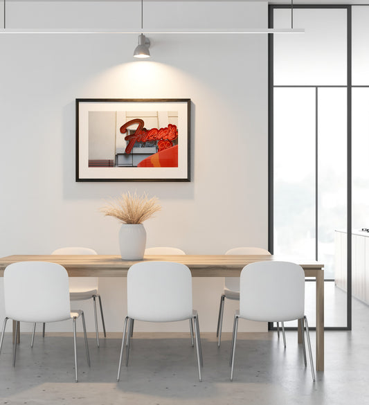 A framed abstract art print featuring a red and orange design, displayed on a white wall above a modern dining table with white chairs in a bright, contemporary dining room.