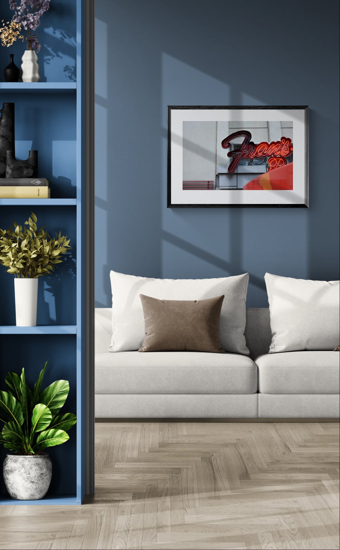 A framed abstract art print with red, black, and white tones hangs on a dark blue wall above a light gray sofa with white and brown pillows. The room includes a blue shelving unit with plants and decor, and a light wood herringbone floor.