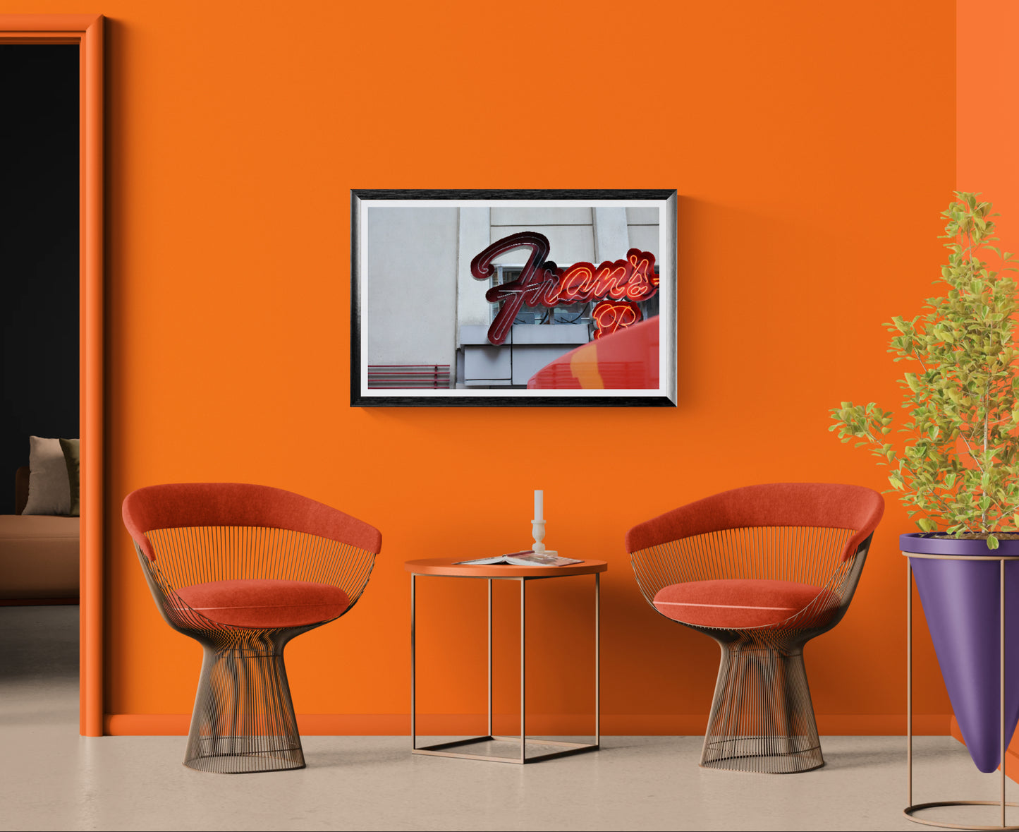 A framed art print featuring a red neon-style 'Frans' sign, displayed on a vibrant orange wall in a modern living room setting with two orange mesh chairs and a small side table.