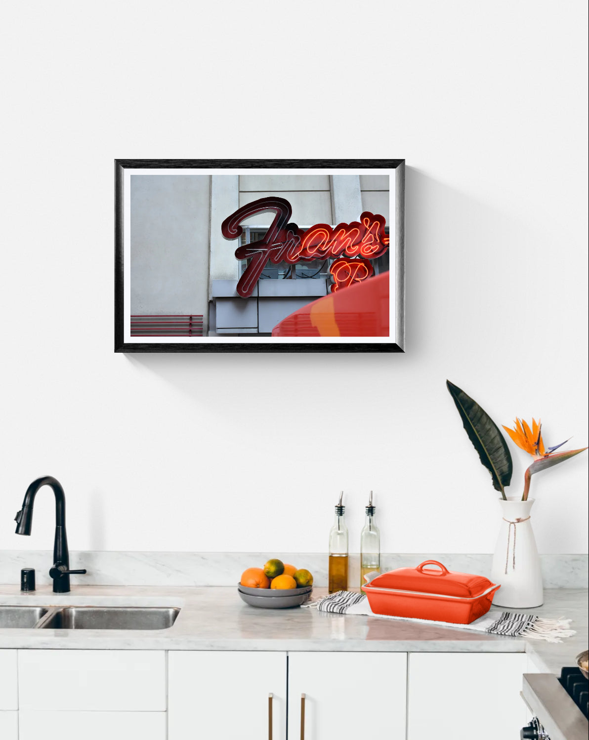 A framed wall art print featuring a red neon-style 'Fran's' sign against a gray building facade, displayed above a kitchen counter with a sink, fruit, bottles, and a bright orange lidded casserole dish.