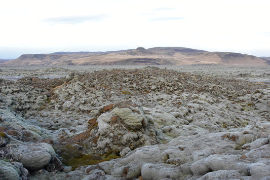 A landscape photograph featuring a field of lava rocks with patches of green moss, with a desolate and rugged terrain in the background.