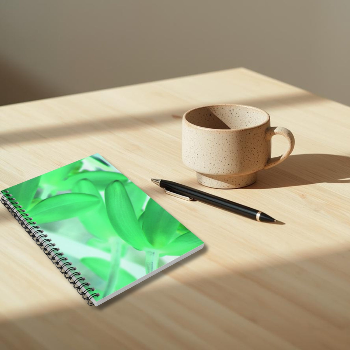 Notebook with a green leaf design, pen, and ceramic cup on a wooden surface.